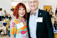 A woman and man pose together at a formal gathering, both smiling and wearing name tags, with a lively event setting behind them.