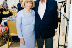 A joyful elderly couple standing close at a social event, surrounded by other attendees and event decorations, capturing a moment of happiness.