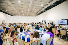A large community dinner event with many guests seated at round tables, enjoying a meal in a spacious hall.