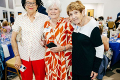 A group of three elderly women enjoying a social event, smiling and standing close together in a well-lit hall with tables and guests in the backgroun.