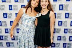 Two girls standing together at a Temple Israel event, smiling and enjoying the occasion. Background features Temple Israel branding.