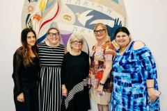 Group of five women posing for a photo at an indoor event with a colorful mural backdrop.
